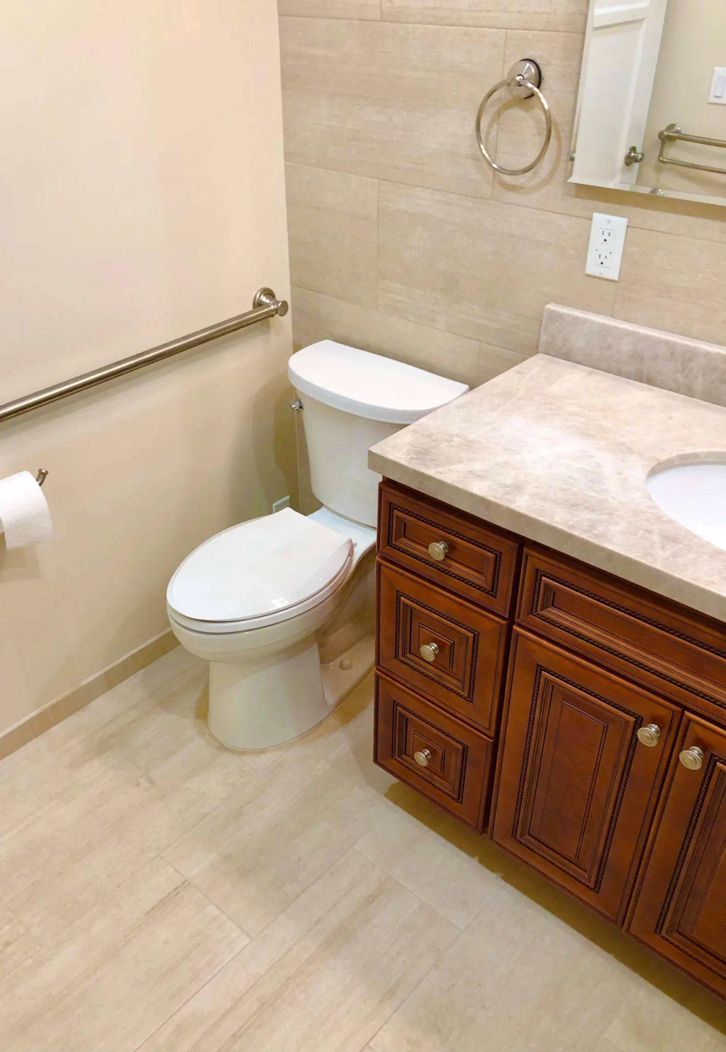 bathroom-remodels-5 Accessible bathroom with a toilet, wooden vanity, grab bar, and toilet paper holder on a tiled floor.