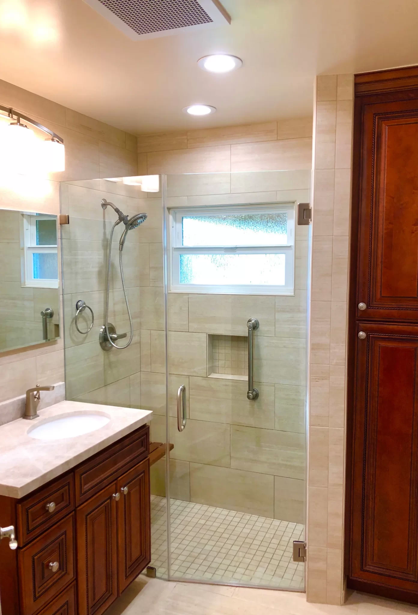 bathroom-remodels-6 Modern bathroom with glass shower, wood cabinets, and beige tile walls, well-lit with ceiling lights.