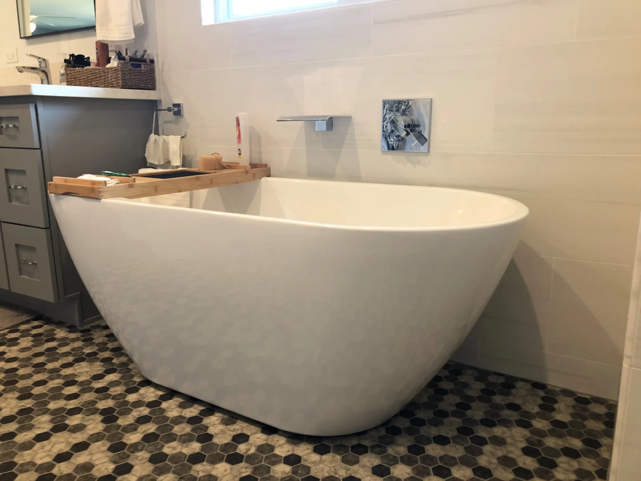 bathroom-remodels-9 Modern white freestanding bathtub on hexagon tile floor, with a wooden bath tray, in a bright bathroom.