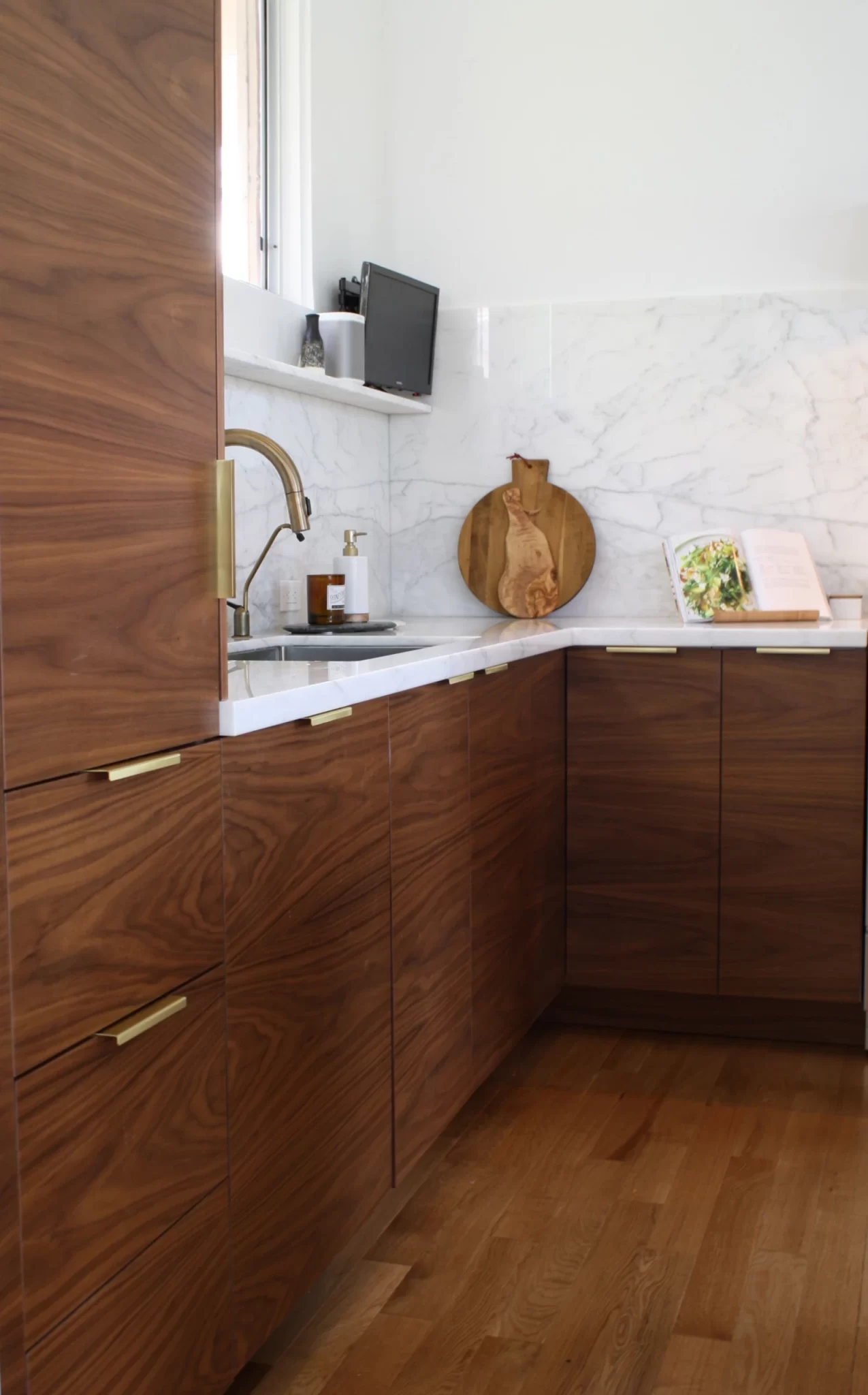 custom-cabinetry-carpentry-1 Modern kitchen with walnut cabinets, marble backsplash, gold faucet, and a cutting board by the sink.