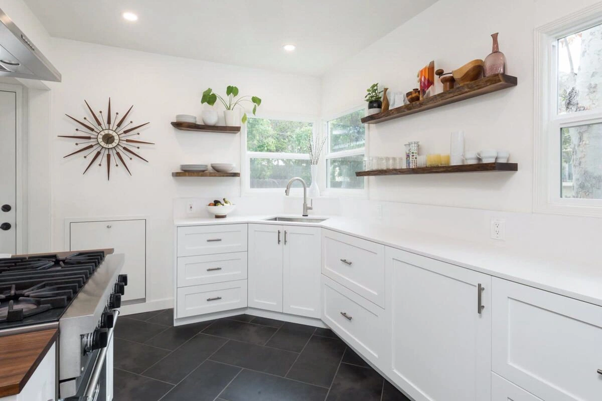 custom-cabinetry-carpentry-16 Modern white kitchen with black tile floor, open wooden shelves, and a starburst clock on the wall.