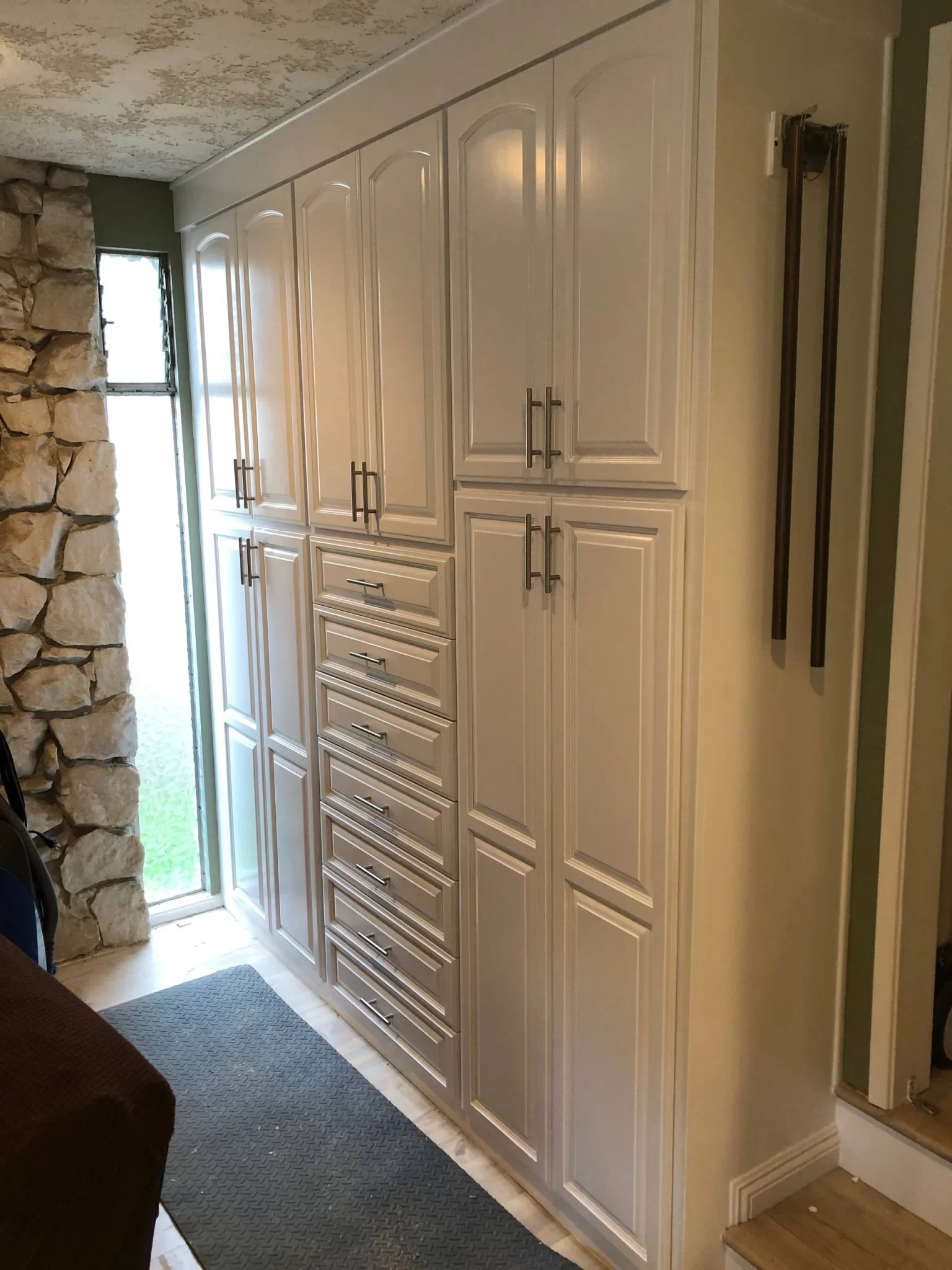custom-cabinetry-carpentry-9 Tall, white built-in cabinets with multiple doors and drawers next to a stone wall and window.