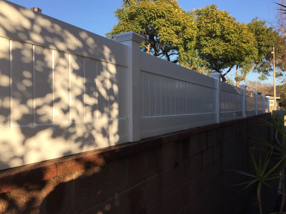 custom-fences-and-gates-1 White vinyl fence on top of a brick wall, with tree shadows and greenery in the background.