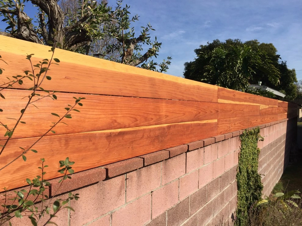 custom-fences-and-gates-3 A brick wall topped with new wooden planks, with plants growing nearby, under a blue sky.