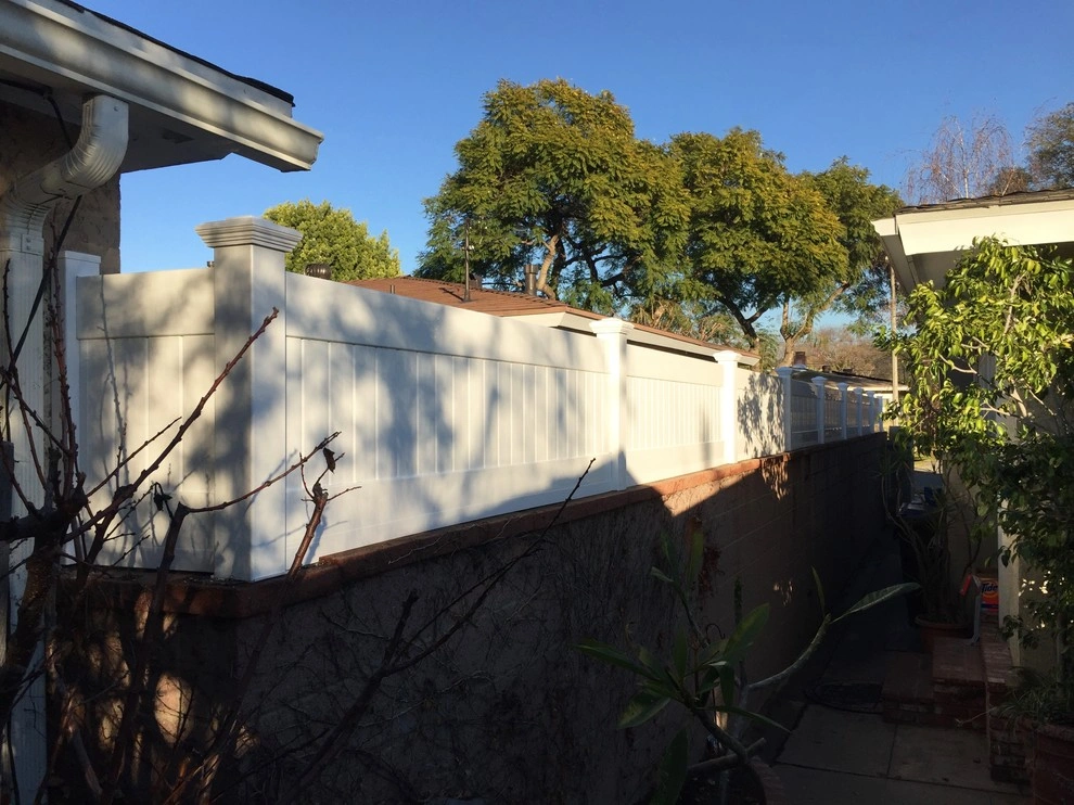 custom-fences-and-gates-4 A tall white vinyl fence on a brick wall separates two houses with trees in the background.