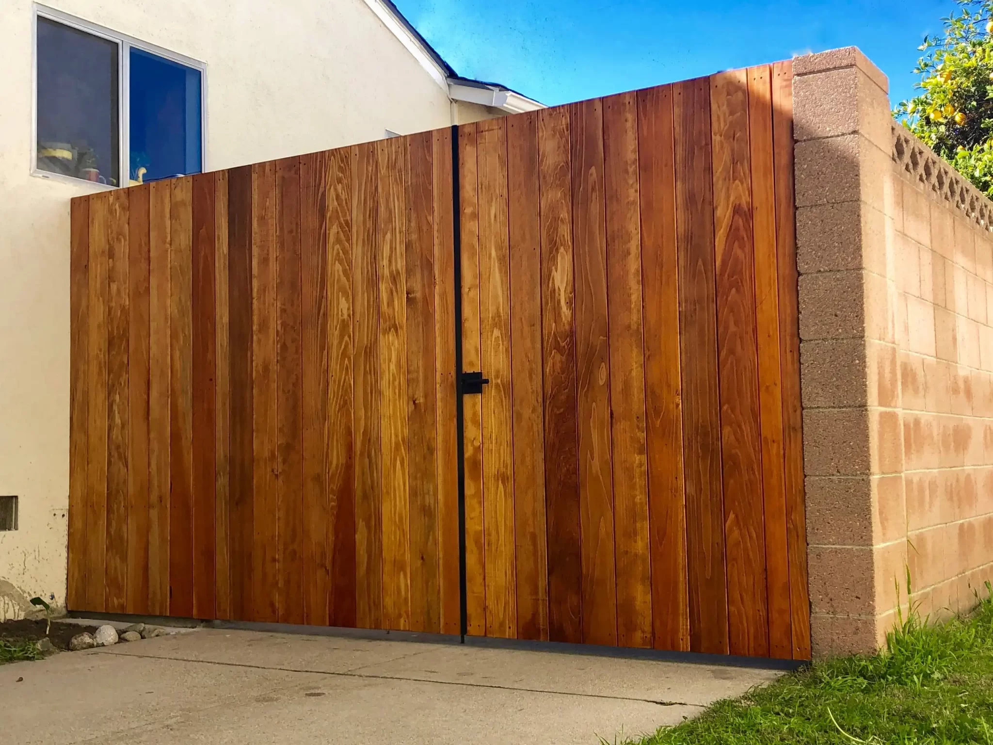 custom-fences-and-gates-7 Tall wooden gate with vertical panels next to a beige brick wall and house, under a blue sky.
