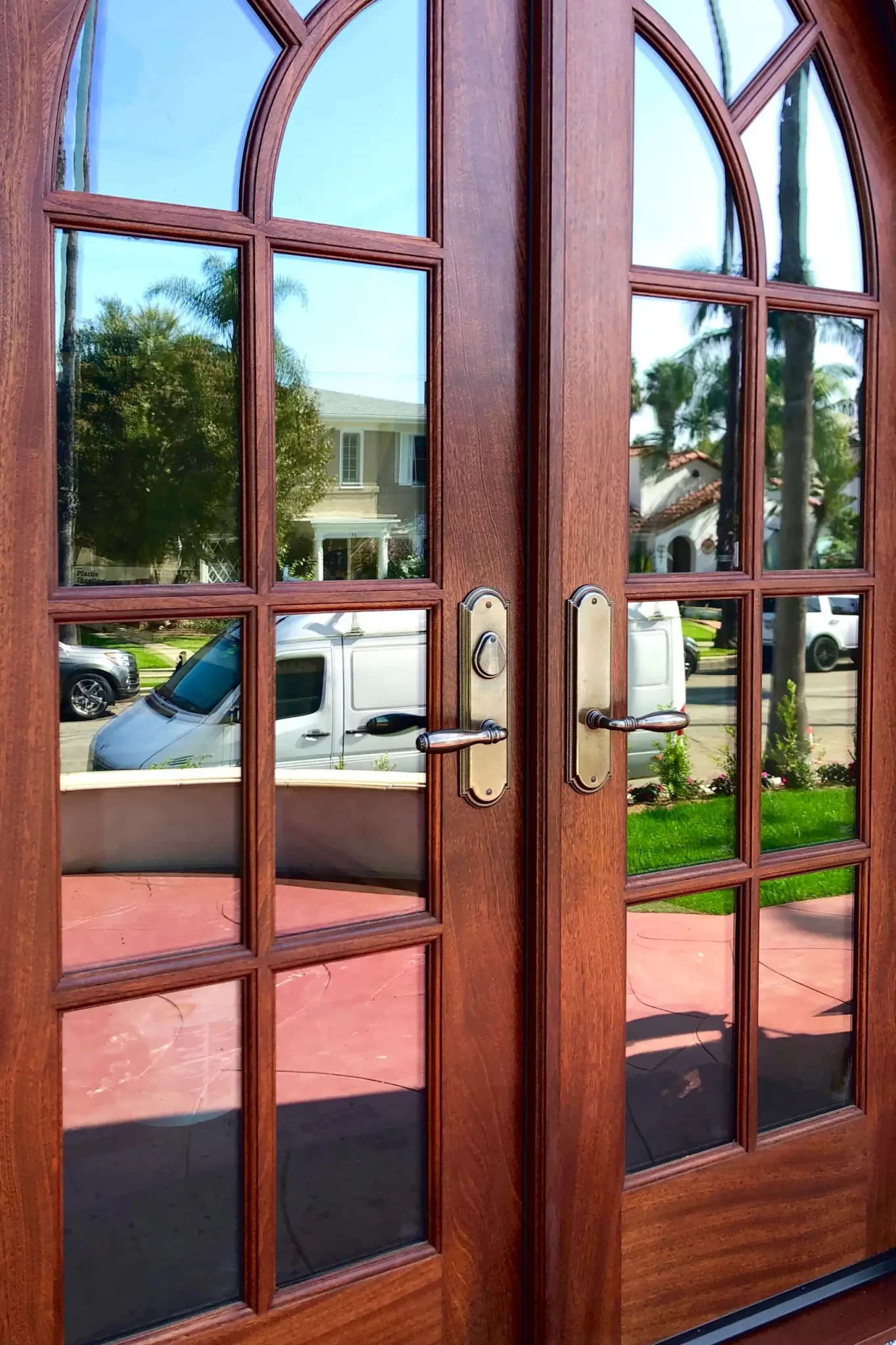 doors-and-windows-1 Double wooden doors with glass panels reflecting houses, trees, and parked cars on a sunny day.