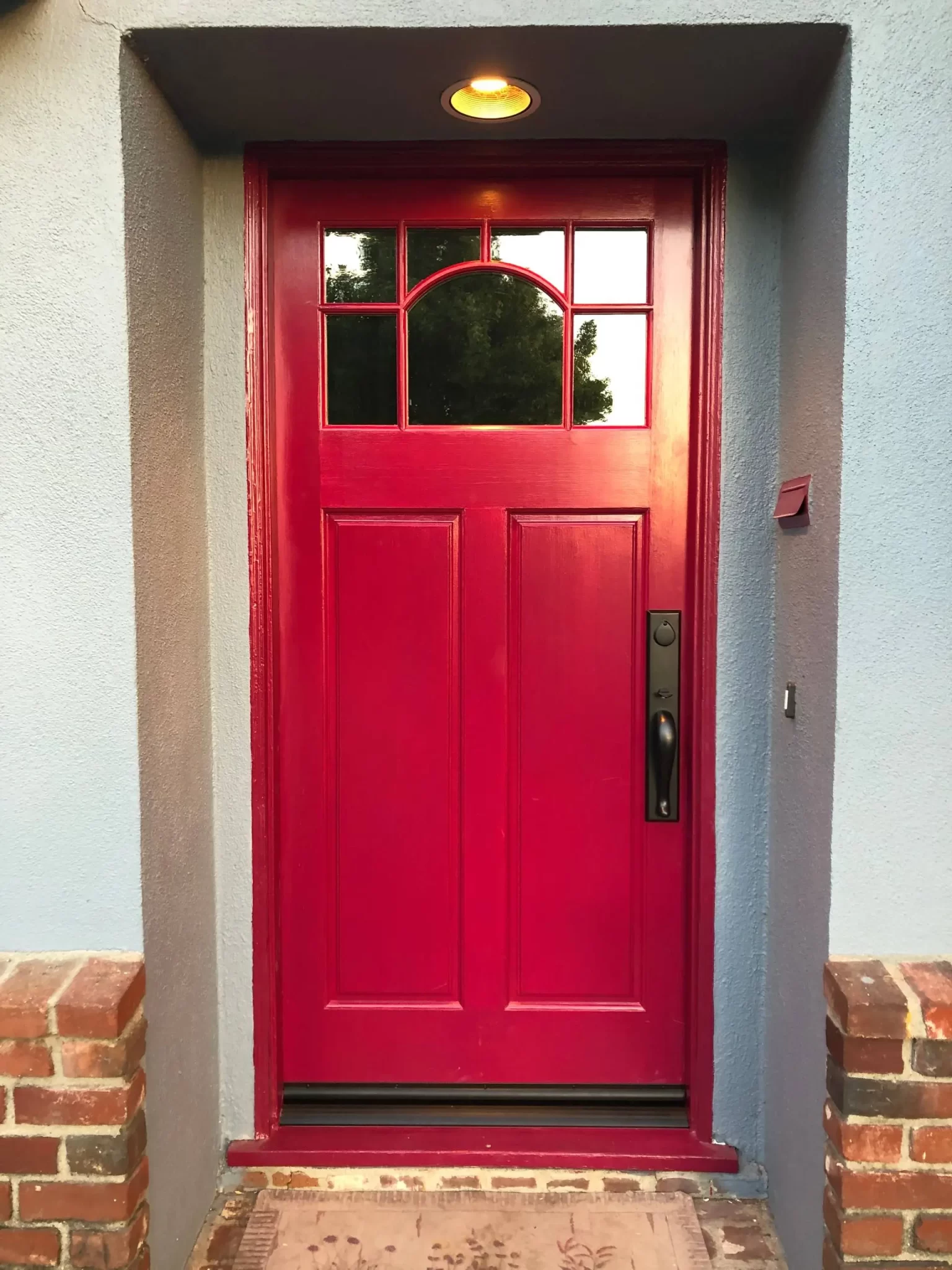 doors-and-windows-4 Bright red front door with windows at the top, set in a blue wall with brick steps and a welcome mat.