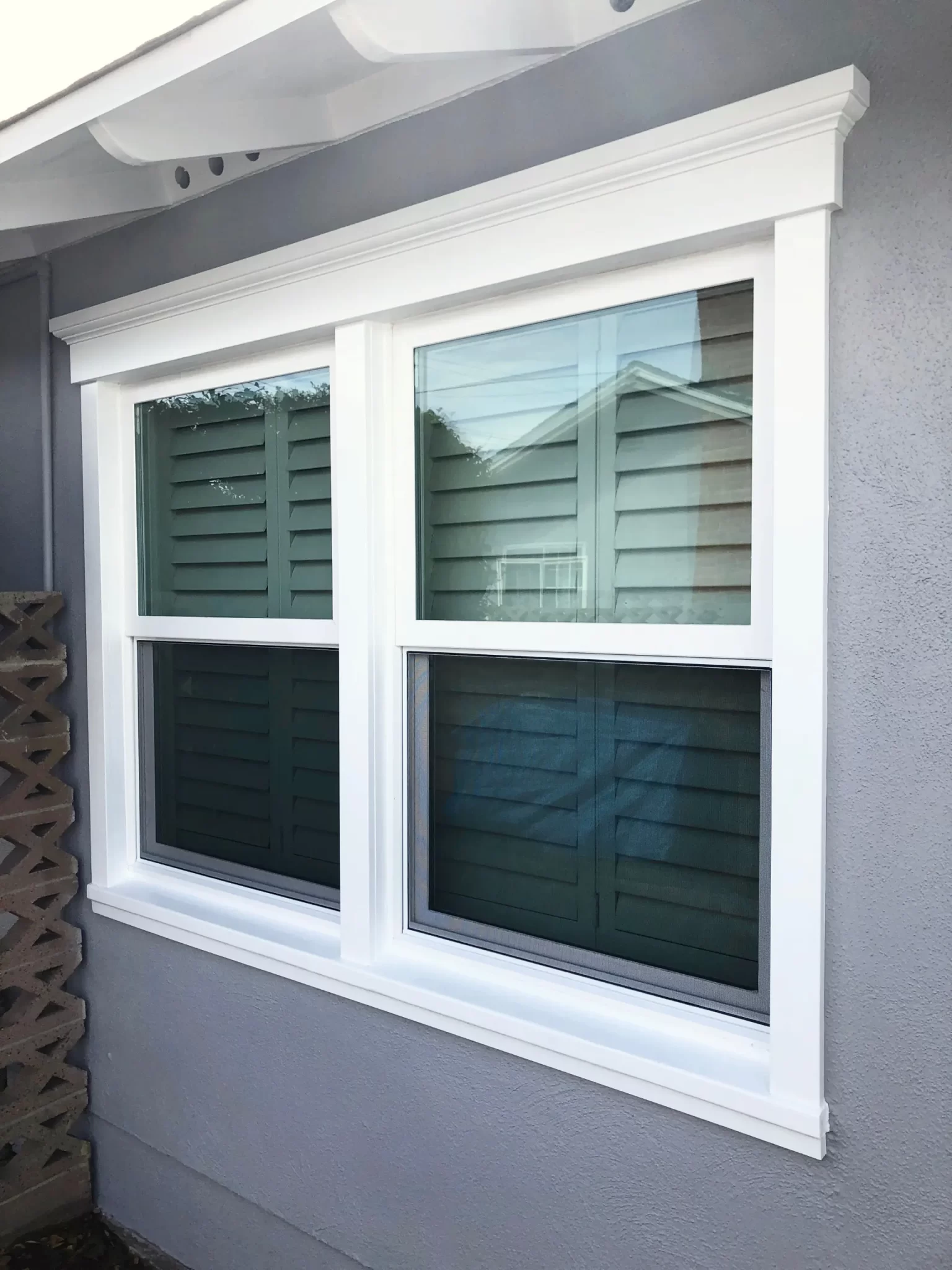 doors-and-windows-8 White-framed double window with shutters on a gray exterior wall, reflecting a nearby house.