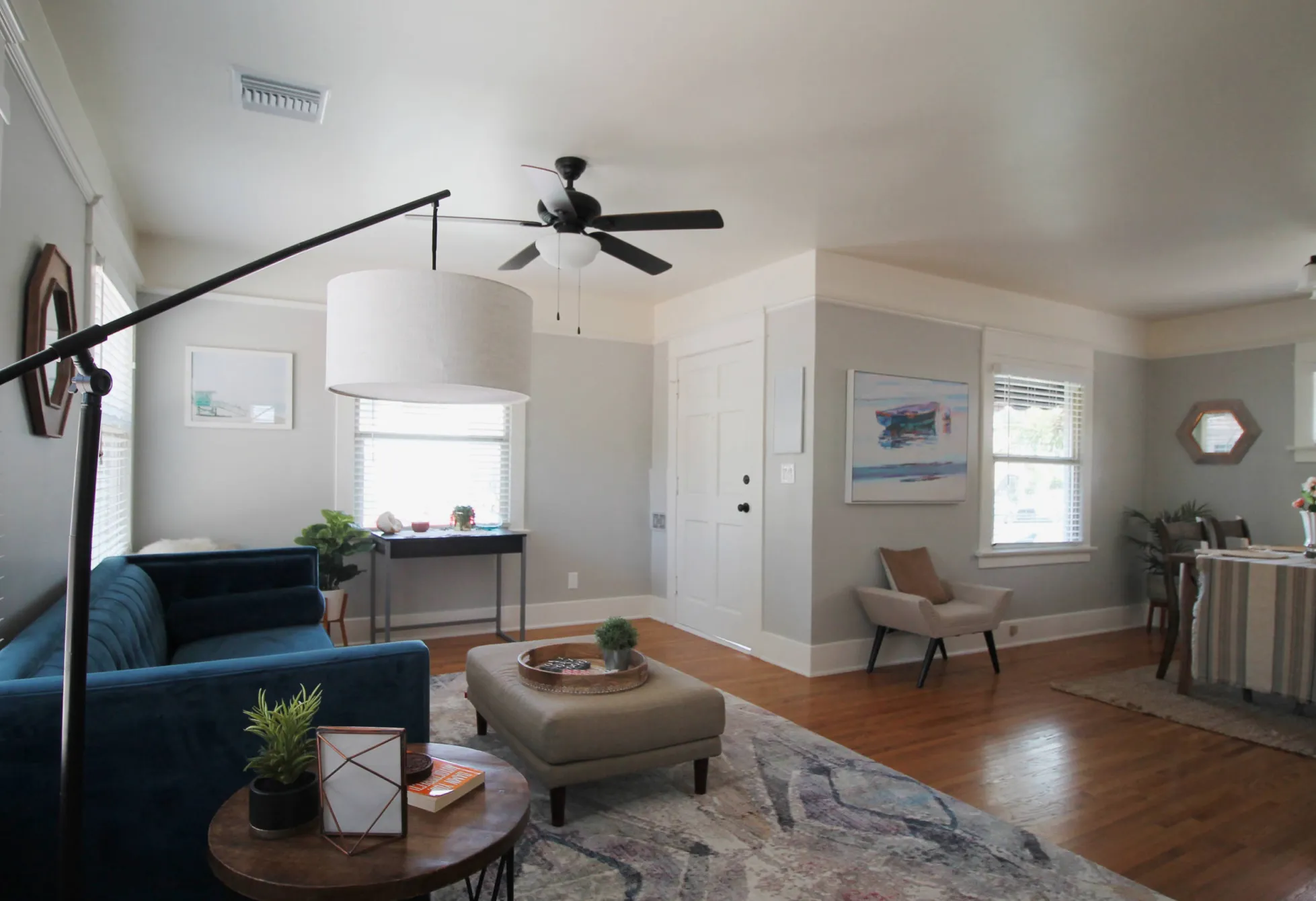 Modern living room with a blue sofa, floor lamp, ceiling fan, and a small table. Neutral walls and hardwood floor.
