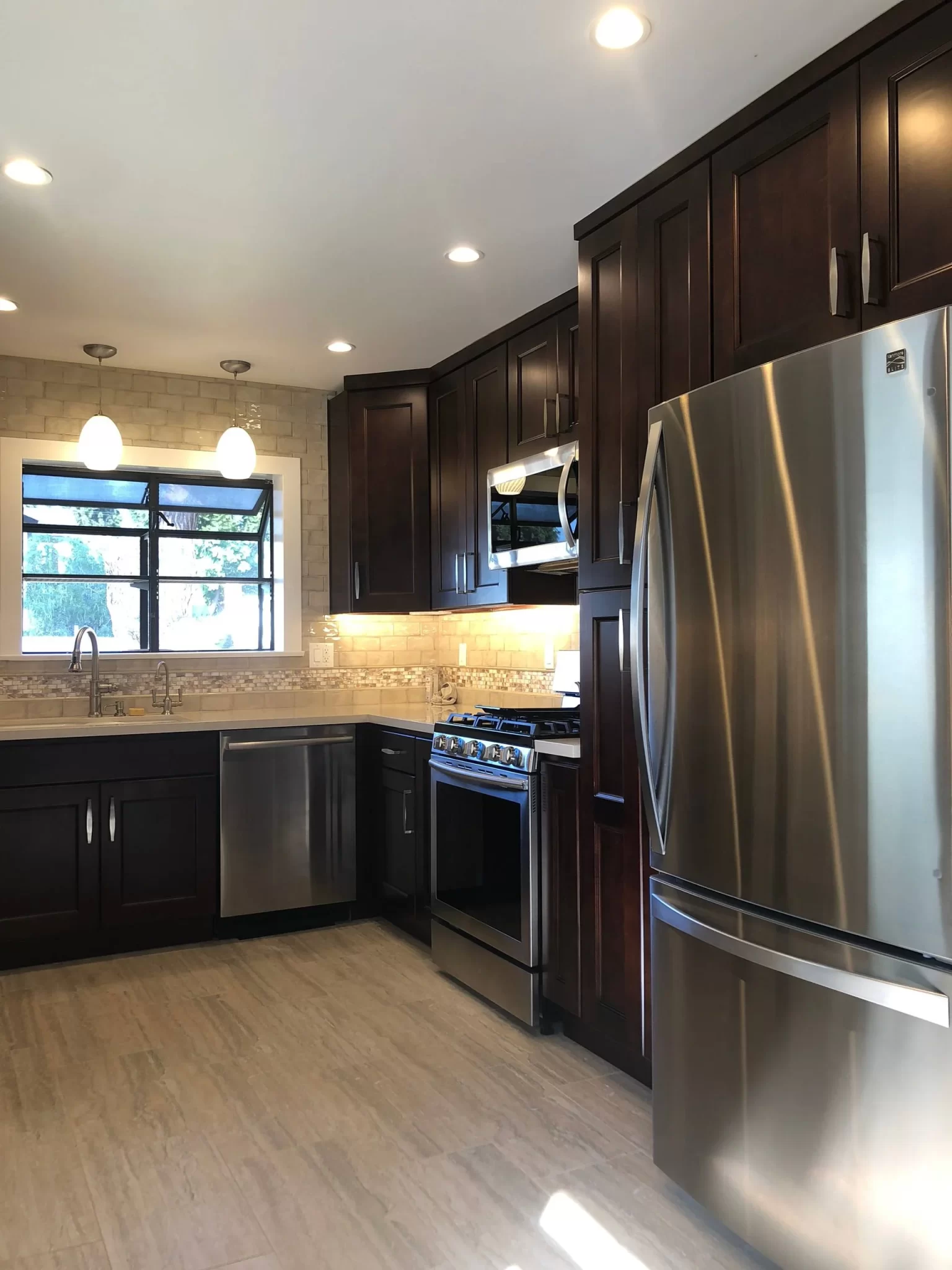 kitchen-remodels-10 Modern kitchen with dark wood cabinets, stainless steel appliances, and light tile floor, lit by recessed lights.