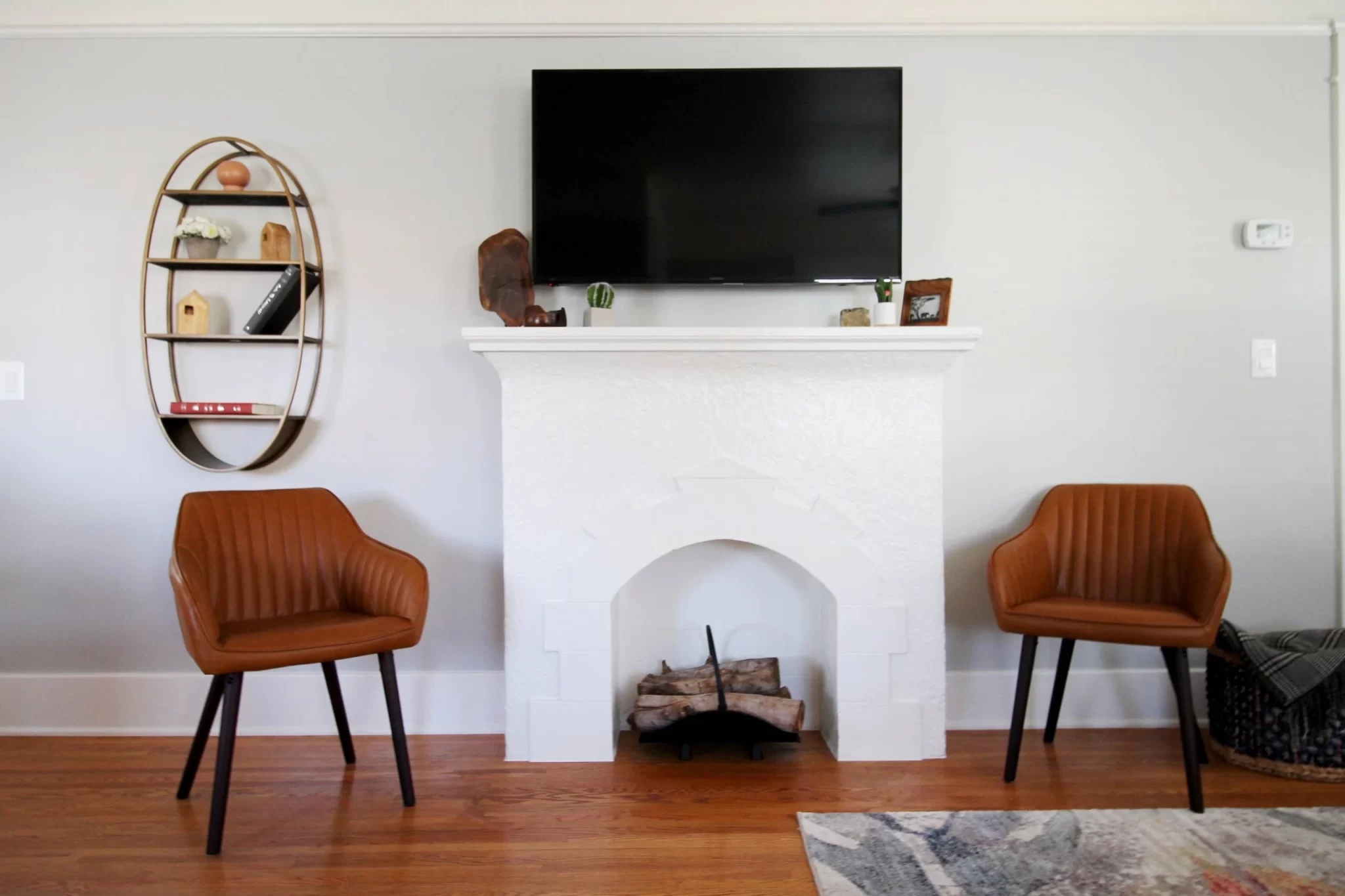 living-spaces-3 Modern living room with two brown chairs, a wall-mounted TV above a white fireplace, and a round bookshelf.