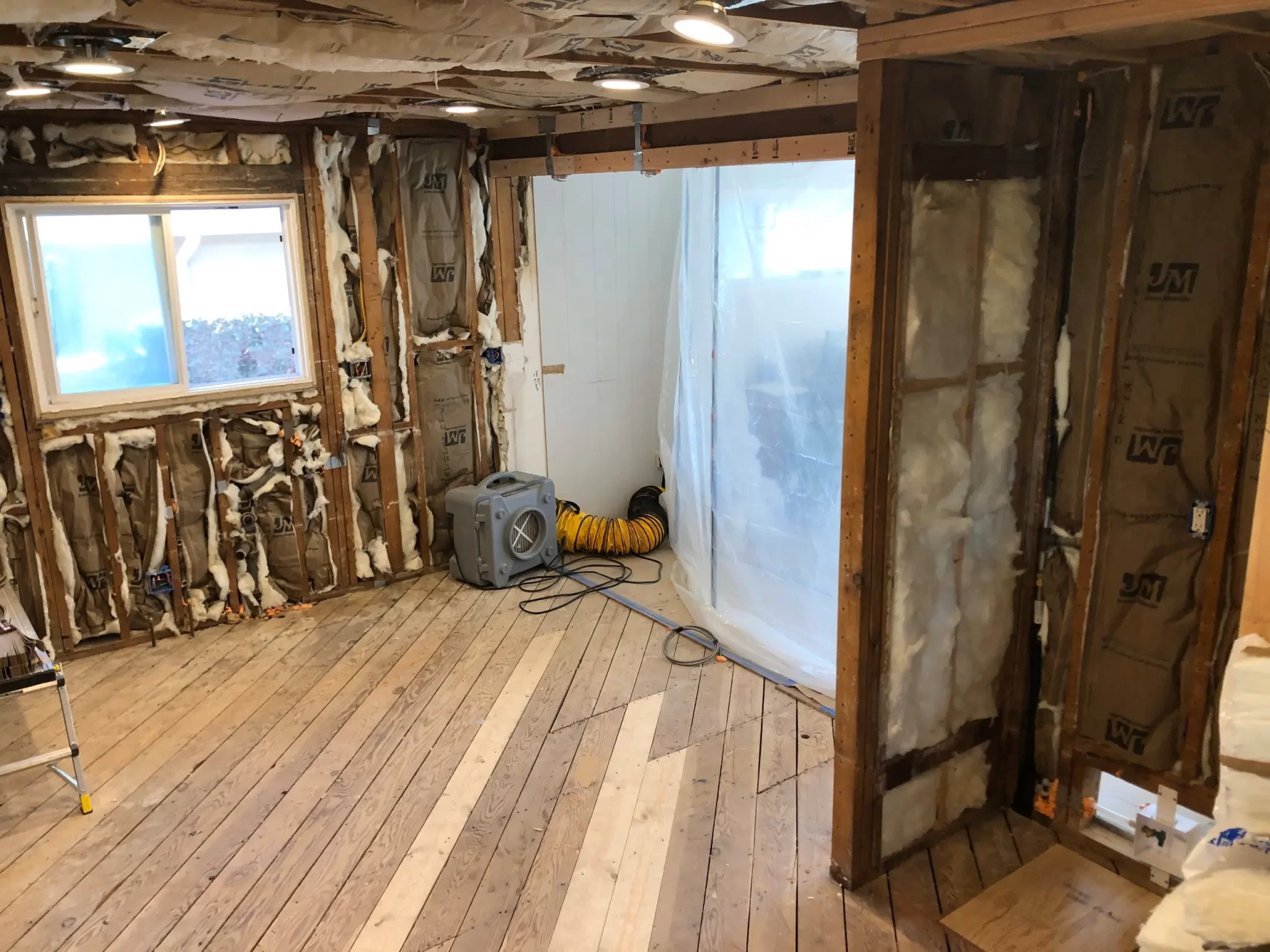 projects-in-process-2 Room under renovation with exposed insulation, wood floors, a window, and construction equipment.
