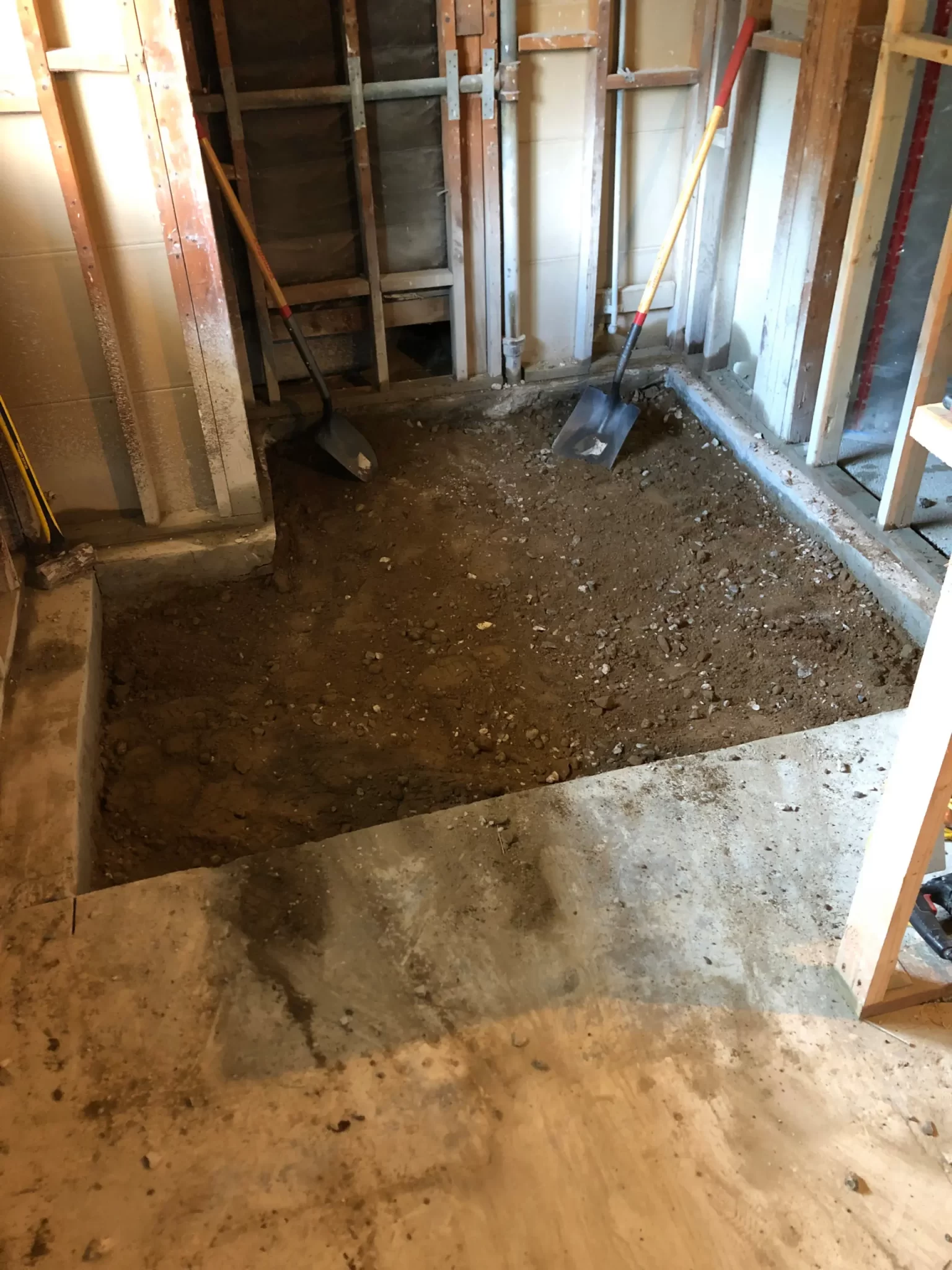 projects-in-process-3 A room under renovation with a dirt-filled area and two shovels propped against exposed wall studs.