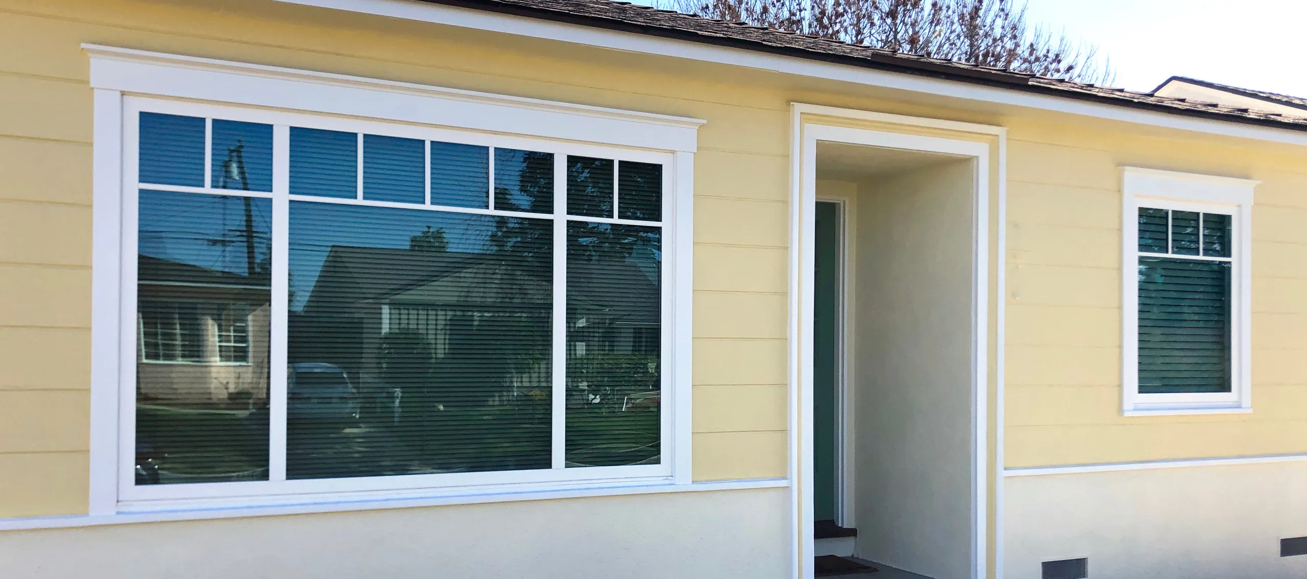 Yellow house with large window reflecting a street view and a recessed entryway with a simple white door frame.