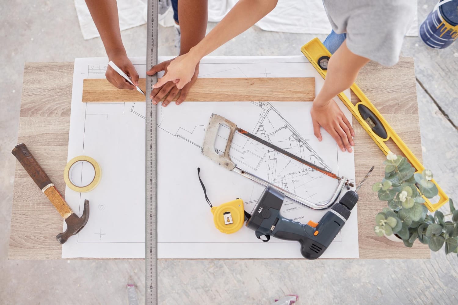 Two people work on blueprints at a table with tools, including a saw, hammer, drill, and measuring tape.