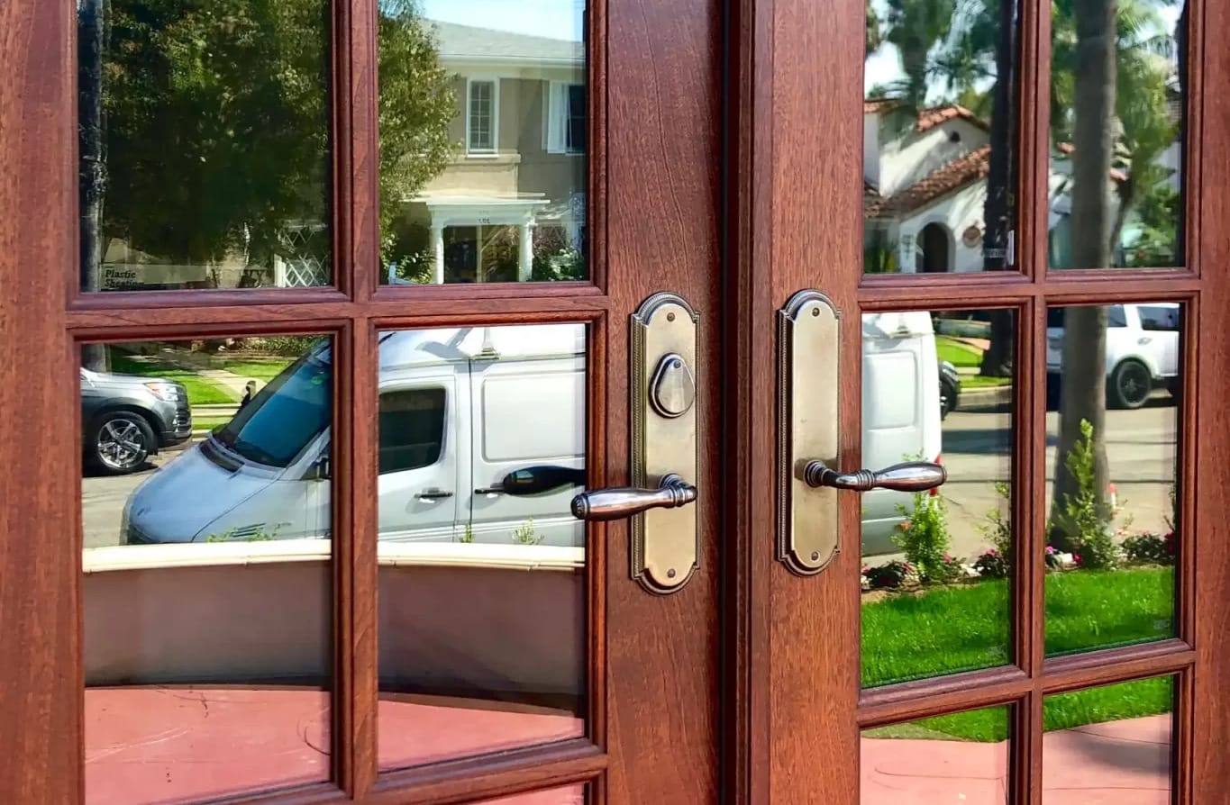 Brown wooden double doors reflect a white van, houses, and greenery on their glass panels.