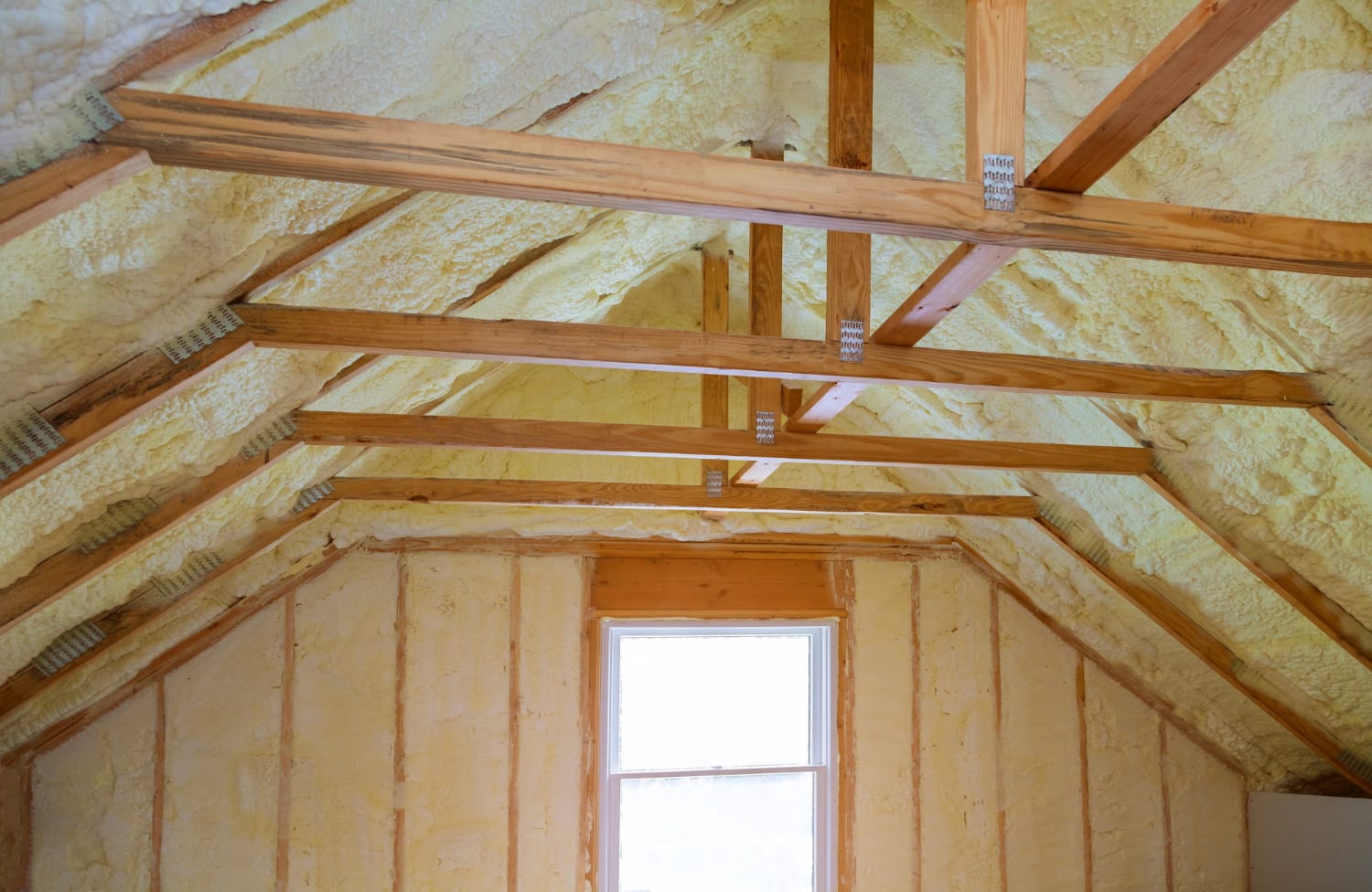 Spray foam insulation on attic ceiling and walls with exposed wooden beams and a window.