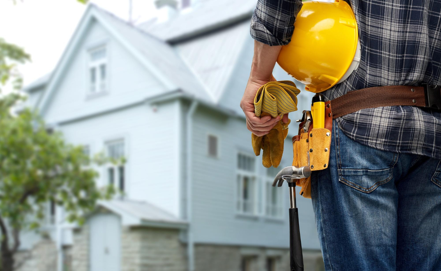 A construction worker holds a yellow hard hat and blueprint, standing in front of a house.